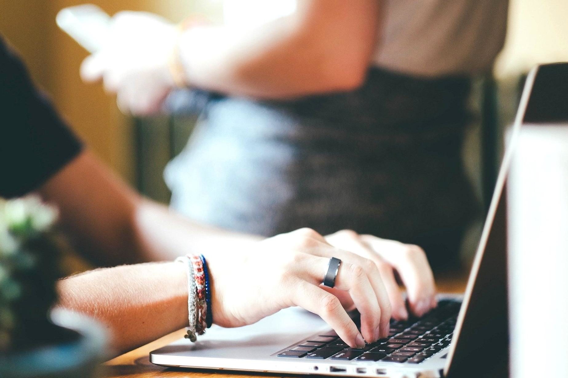 Typing on a laptop computer. A person stands in the background.